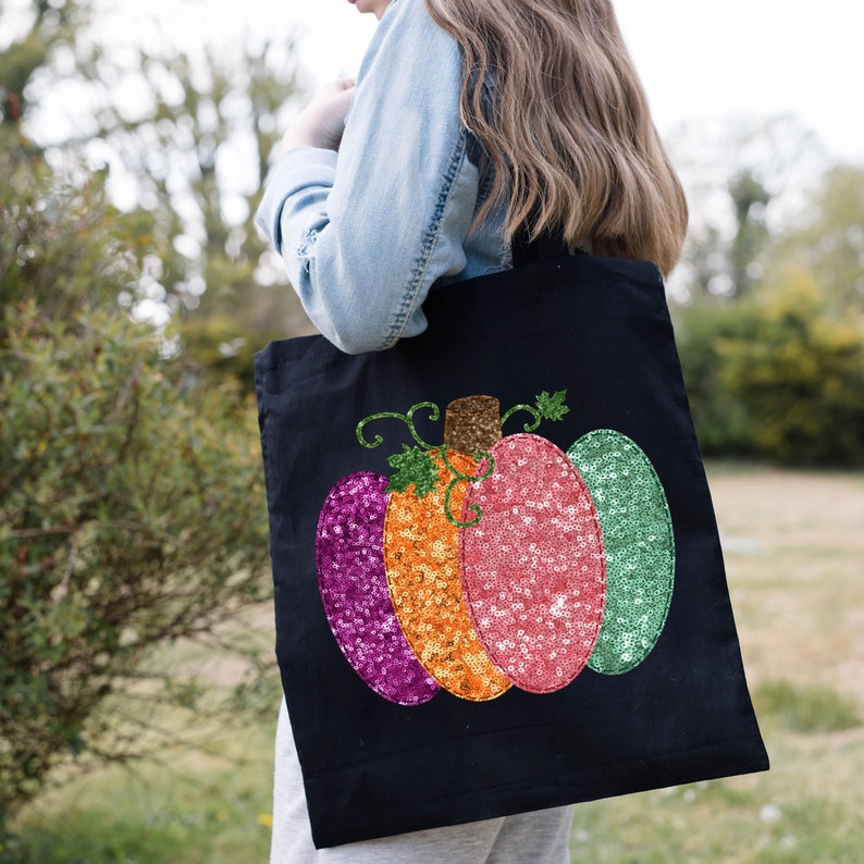 May include: A black tote bag with a sequined pumpkin design. The pumpkin is made of pink, orange, and green sequins.