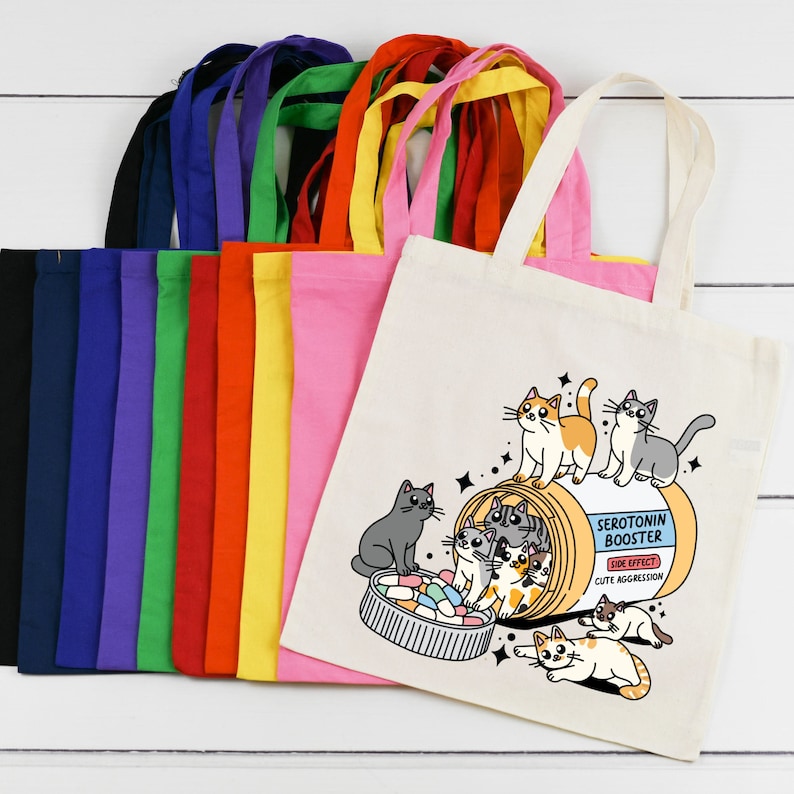 May include: A set of 12 colorful tote bags with a white tote bag featuring a cartoon illustration of cats in a pill bottle with the text "Serotonin Booster" and "Side Effect: Cute Aggression".