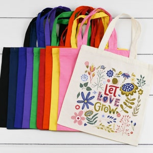 May include: A set of 12 colorful tote bags with a white tote bag featuring a floral design and the text "Let Love Grow".
