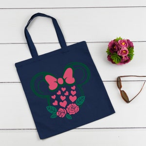 Puede incluir: Una bolsa de tela azul marino con una silueta verde y rosa de un personaje de dibujos animados con un lazo. El personaje est&aacute; rodeado de corazones rosas y hojas verdes con rosas rosas.