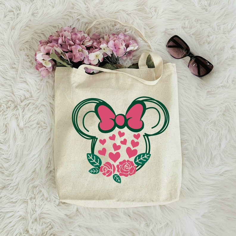 Puede incluir: Una bolsa de lona blanca con una silueta verde y rosa de una cabeza de rat&oacute;n con un lazo y corazones. La cabeza del rat&oacute;n est&aacute; rodeada de hojas verdes y rosas rosas.