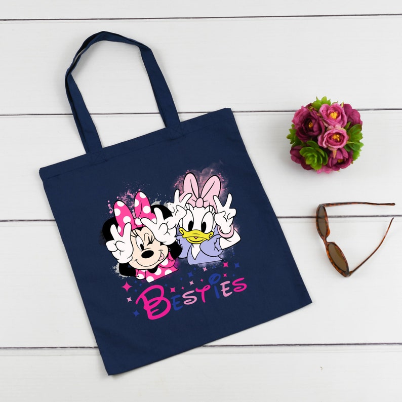 May include: A navy blue tote bag featuring a pink and white graphic of Minnie Mouse and Daisy Duck. The graphic shows the characters with their hands raised in a peace sign. The text "Bestes" is printed below the characters.