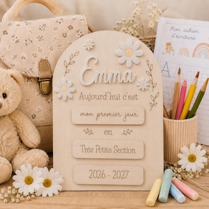 Peut inclure: Une plaque en bois arquée avec le prénom Emma en relief blanc, décorée de marguerites blanches et du texte gravé Aujourd'hui c'est mon premier jour en Très Petite Section 2026-2027, entourée d'un ours en peluche et de crayons de couleur.