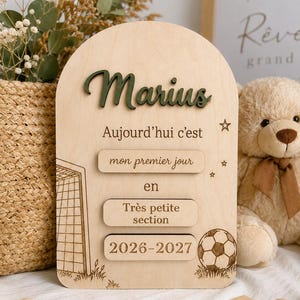 Peut inclure: Une plaque en bois en forme d'arche avec le prénom Marius en relief vert foncé. Le texte indique "Aujourd'hui c'est mon premier jour en Très petite section 2026-2027". Elle comporte des gravures d'un but de football, d'un ballon et d'étoiles.