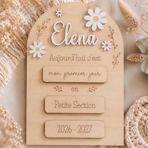 Pode incluir: Uma placa de madeira em arco com o nome Elena em letras brancas em relevo, acompanhada por flores brancas recortadas e o texto gravado: Aujourd'hui c'est mon premier jour en Petite Section 2026-2027, decorada com motivos de folhas.