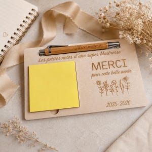 Peut inclure: Un support à notes rectangulaire en bois avec un stylo en bambou, un bloc-notes adhésif jaune carré et des gravures laser indiquant "Les petites notes d'une super Maitresse", "MERCI pour cette belle année" et "2025-2026" avec des motifs floraux.