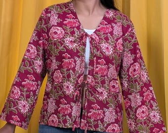 May include: A long-sleeved jacket in a deep magenta hue featuring a repeating floral pattern in shades of pink, white, and olive green. The garment has a V-neckline and two sets of thin fabric ties at the center front for closure.