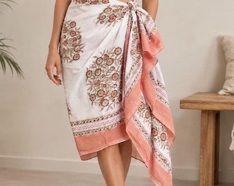 May include: A white sarong wrap skirt featuring a repeating floral pattern in shades of brown and green, finished with a wide coral-coloured border along the edges. The fabric is tied at the waist, creating a draped effect with an asymmetrical hemline.
