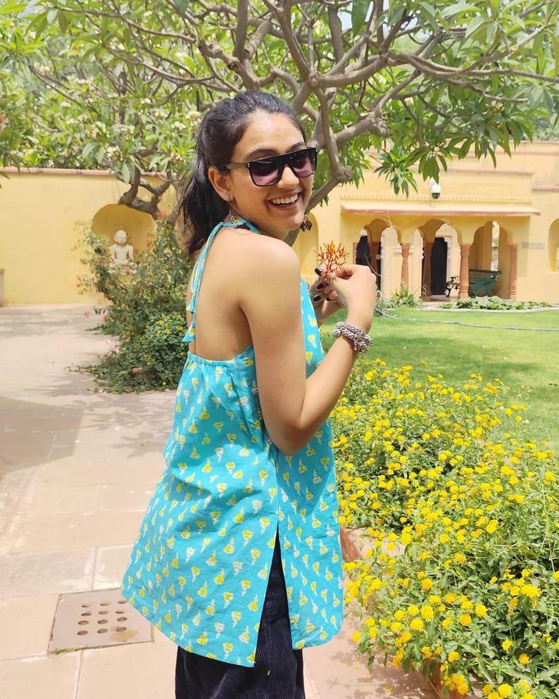 May include: A woman wearing a blue and yellow floral print halter top with a side slit. She is smiling and holding a small flower in her hand.