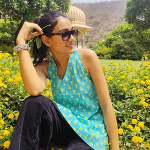 May include: A woman wearing a blue and yellow floral print sleeveless top and black trousers sits in a garden setting. She is wearing sunglasses and has her hair pulled back.