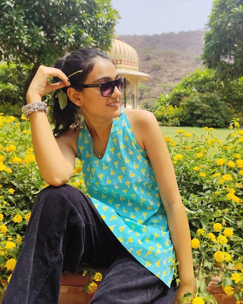 May include: A woman wearing a blue and yellow floral print sleeveless top and black trousers sits in a garden setting. She is wearing sunglasses and has her hair pulled back.