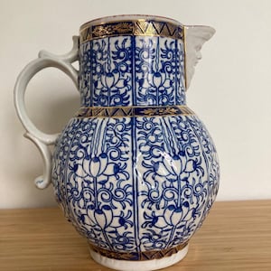 May include: A blue and white ceramic jug with a gold rim and handle. The jug features a floral pattern and a small face on the spout. The handle is curved and the jug sits on a wooden surface.