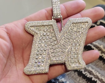 May include: A silver-colored pendant in the shape of the letter "M", encrusted with numerous small, clear gemstones. The pendant has a rectangular bail at the top for a chain. The gemstones are set closely together, creating a sparkling effect.