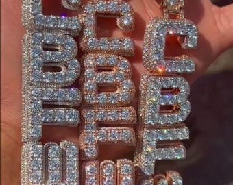 May include: Three iced-out pendant necklaces spelling out the word "CBME". The pendants are encrusted with sparkling, clear gemstones. One is silver-toned, and two are rose gold-toned. The necklaces are displayed against a blurred background.