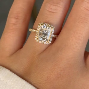 May include: A gold engagement ring with a large, rectangular, clear diamond center stone surrounded by a halo of smaller diamonds. The ring is set on a thin gold band.