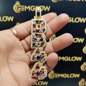 May include: Gold pendant necklace with the number 24 in large, bold letters. The letters are encrusted with colorful gemstones, including pink, red, blue, and clear stones.