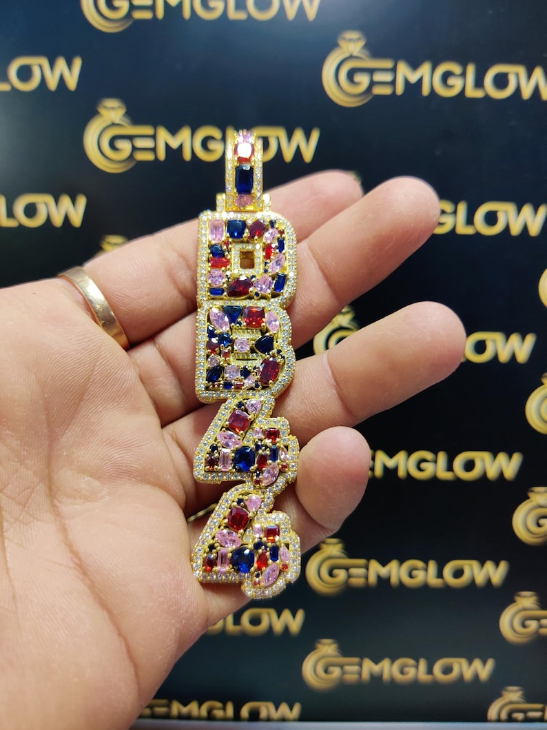 May include: Gold pendant necklace with the number 24 in large, bold letters. The letters are encrusted with colorful gemstones, including pink, red, blue, and clear stones.