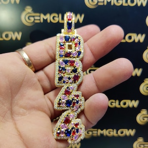 May include: Gold pendant with the letters "BR4" set with colorful gemstones. The pendant is held in a hand.