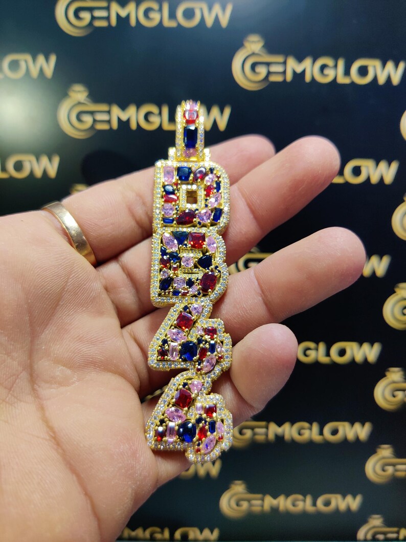 May include: Gold pendant with the letters "BR4" set with colorful gemstones. The pendant is held in a hand.