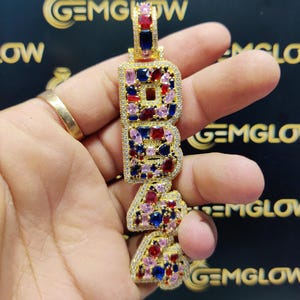 May include: Gold pendant necklace with the letters "B.R.A." spelled out in large, bold letters. Each letter is encrusted with colorful gemstones, including red, blue, and pink.