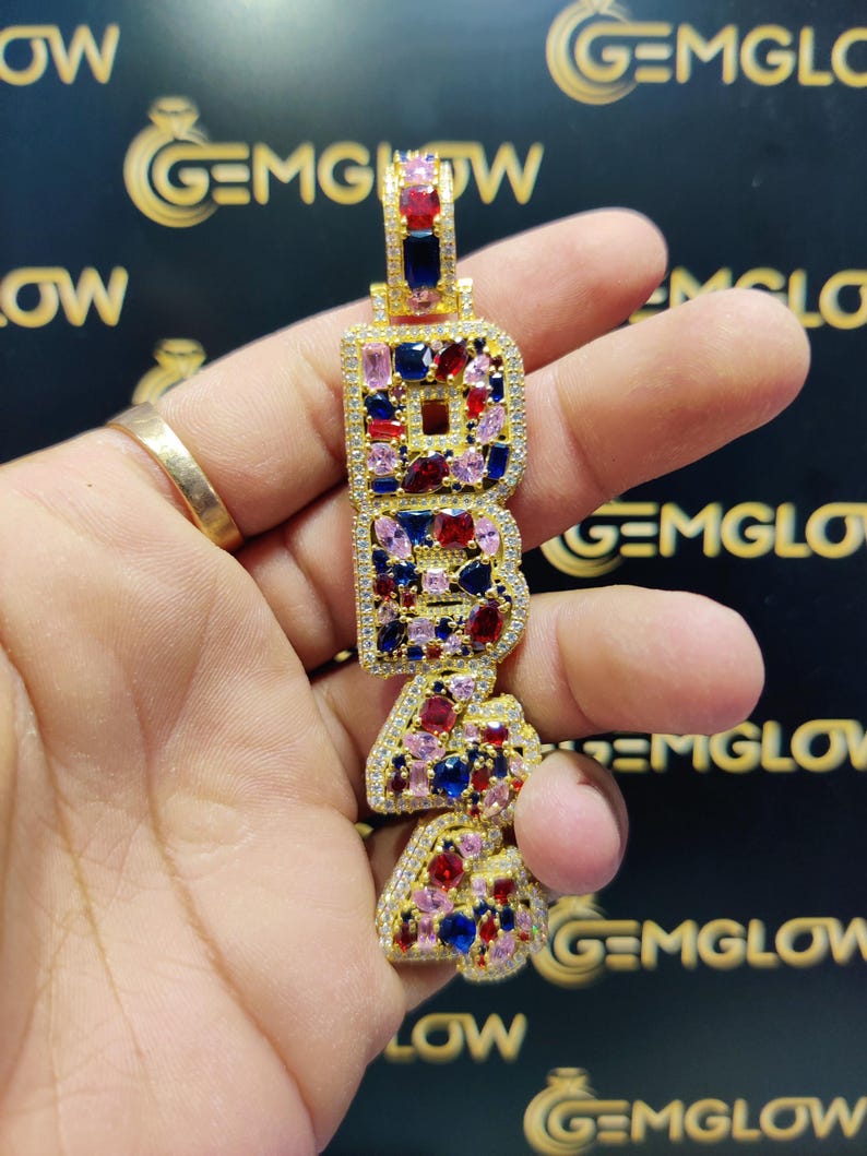 May include: Gold pendant necklace with the letters "B.R.A." spelled out in large, bold letters. Each letter is encrusted with colorful gemstones, including red, blue, and pink.