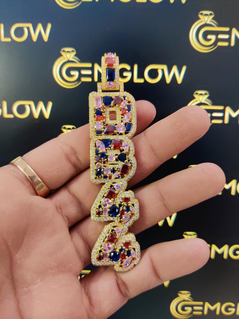 May include: Gold pendant with the letters "1942" set with colorful gemstones, including red, blue, and pink.