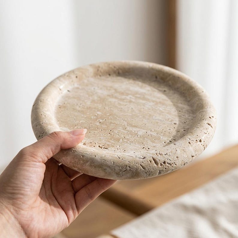 May include: A round, beige stone tray with a slightly raised edge. The tray is held by a hand, showcasing its natural texture and color variations. The tray's diameter is approximately 6 inches.