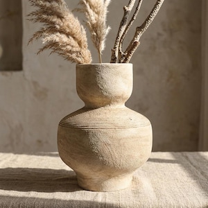 Op de afbeelding: Een beige, getextureerde vaas met een bolvormige basis en een smallere hals, met gedroogd pampasgras en takjes. De vaas heeft horizontale geribbelde details rond het midden. De neutrale tinten en natuurlijke elementen creëren een rustieke, bohemien esthetiek.