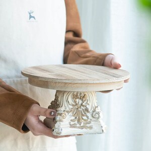 以下が含まれることがあります： 自然な明るい茶色の木目調の天板を持つ円形の木製ケーキスタンド。ベース部分はオフホワイトに塗装された素材で作られており、装飾的な彫刻のレリーフと、使い古されたような質感のゴールドのアクセントが施されています。