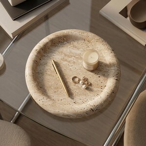 May include: A beige, round tray with a textured surface holds a gold pen, a small candle, and several pieces of jewelry. The tray sits on a glass table, with other items visible in the background.