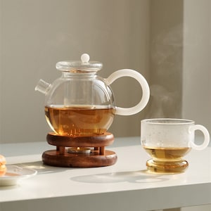 Puede incluir: Una tetera de vidrio transparente con asa blanca se encuentra sobre un soporte de madera. La tetera está llena de té y tiene un filtro integrado. Una taza de té de vidrio transparente con asa está llena de té y se encuentra sobre una superficie blanca.