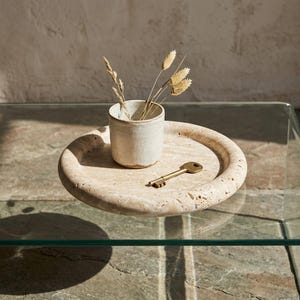 May include: A beige stone tray with a small, off-white ceramic cup holding dried wheat stalks. A brass key rests on the tray's surface. The tray sits on a glass table, creating a minimalist, natural aesthetic.