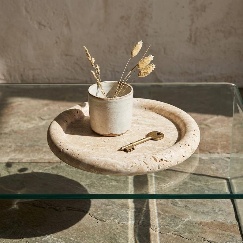 May include: A beige stone tray with a small, off-white ceramic cup holding dried wheat stalks. A brass key rests on the tray's surface. The tray sits on a glass table, creating a minimalist, natural aesthetic.