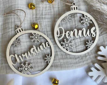 Könnte beinhalten: Zwei kreisförmige Ornamente aus hellem Holz mit ausgeschnittenen Schneeflockenmotiven und den Namen Christine und Dennis in Schreibschrift. Jedes Ornament hat eine Schlaufe mit Bindfaden und ist mit mehreren kleinen goldenen Glöckchen versehen.