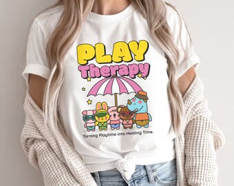 May include: White t-shirt with a graphic of a pink umbrella with the text "PLAY Therapy" in yellow letters.  Under the umbrella are five cartoon animals wearing sunglasses and holding beach towels. The text "Turning Playtime into Healing Time." is below the graphic.