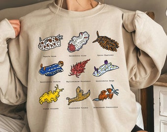 May include: A beige sweatshirt with a graphic print of various colorful sea slugs. The names of each sea slug are printed below the image. The text includes: Vaquita Dai, Chronodoris Albus, Cyerce Figricans, Hypselodoris Festiva, Phyllidiellum Peindinai, Hypselodoris Kanga, Flabellina Crispata, Croslandis Viridis, and Hermissenda Crassicornis.