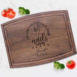 May include: A brown wooden cutting board with a curved edge and a white engraved design that says "Life isn't perfect but your nails can be". The design also includes a hand with a manicured fingernail, stars, and hearts. The text "Jennifer Nail Studio" is engraved below the design.
