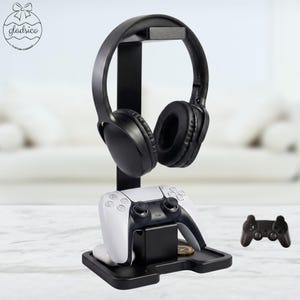 May include: Black headphone stand with a white and black game controller and a black controller. The stand holds a pair of black headphones. The base of the stand has a small tray for accessories.