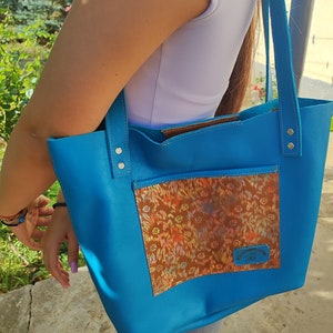 May include: A bright blue leather tote bag with a brown fabric pocket featuring a floral pattern. The bag has two leather straps and a small leather tag with the text "Handmade with Love".