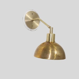 May include: A brass wall sconce with a round, dome-shaped shade. The sconce has a single arm that extends from the wall mount.