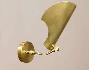 May include: A brass wall sconce with a fan-shaped shade. The sconce has a circular base, a curved arm, and a pivoting shade. The finish is a warm, metallic gold. The design is modern and minimalist.