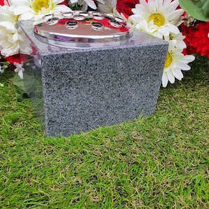 May include: A square grey granite vase with a silver metal insert for holding flowers. The vase is sitting on a bed of green grass.