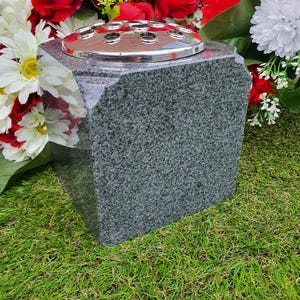 May include: A grey granite vase with a silver metal insert for fresh flowers. The vase is sitting on a bed of green grass.