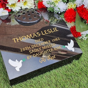 May include: A black granite memorial stone with gold lettering and two white dove decals. The inscription reads "THOMAS LESLIE DIED 26TH DECEMBER 1982 SADLY MISSED BY DAUGHTER KIM At Rest". A flower vase is on top.