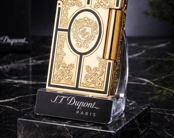 May include: A gold-colored S.T. Dupont lighter with intricate engraved floral patterns and black accents. The lighter is displayed on a black marble base with the brand name "S.T. Dupont Paris" in white text.