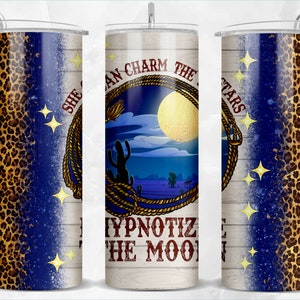 May include: A stainless steel tumbler with a white background and a blue and gold design. The design features a lasso, a cactus, a moon, and stars. The text "She Can Charm The Stars And Hypnotize The Moon" is written on the tumbler.