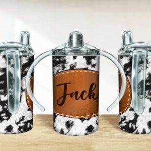 May include: Three clear plastic cups with handles and lids. The cups are decorated with a black and white cow print pattern. The middle cup has a brown leather patch with the name "Jack" written in black.