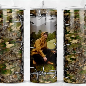 May include: A stainless steel tumbler with a camouflage pattern and barbed wire design. The tumbler has a person's image in the center of the design.