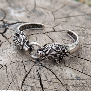 Könnte beinhalten: Ein silberfarbener Armreif aus Metall mit zwei detaillierten Wolfsköpfen, die einander zugewandt sind. Die Wolfsköpfe sind durch einen kreisförmigen Metallring in der Mitte der Öffnung verbunden. Das Metall hat eine polierte Oberfläche mit dunklen, oxidierten Akzenten in den vertieften Bereichen der Fellstruktur.