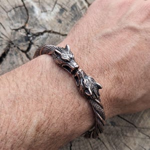 May include: A metallic bracelet featuring two detailed wolf head terminals facing each other. The band consists of a twisted, rope-like design with a dark, oxidized finish that highlights the intricate fur textures and facial features of the wolves.